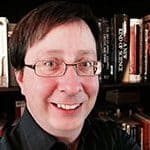 Eine lächelnde Person mit Brille und kurzen dunklen Haaren, die ein dunkles Hemd trägt, steht vor einem Bücherregal voller Bücher.