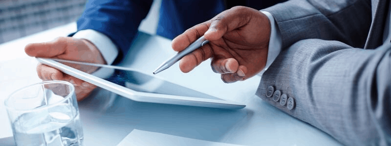 Deux personnes en costume discutent des tests A/B tout en utilisant une tablette. L'une tient la tablette, tandis que l'autre pointe l'écran avec un stylet. Un verre d'eau est posé sur la table à proximité.