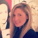 Eine Frau mit langen blonden Haaren lächelt in die Kamera. Sie steht in einem Raum vor einer weißen Orchidee, im Hintergrund ist ein farbenfrohes Porträt einer Frau zu sehen.