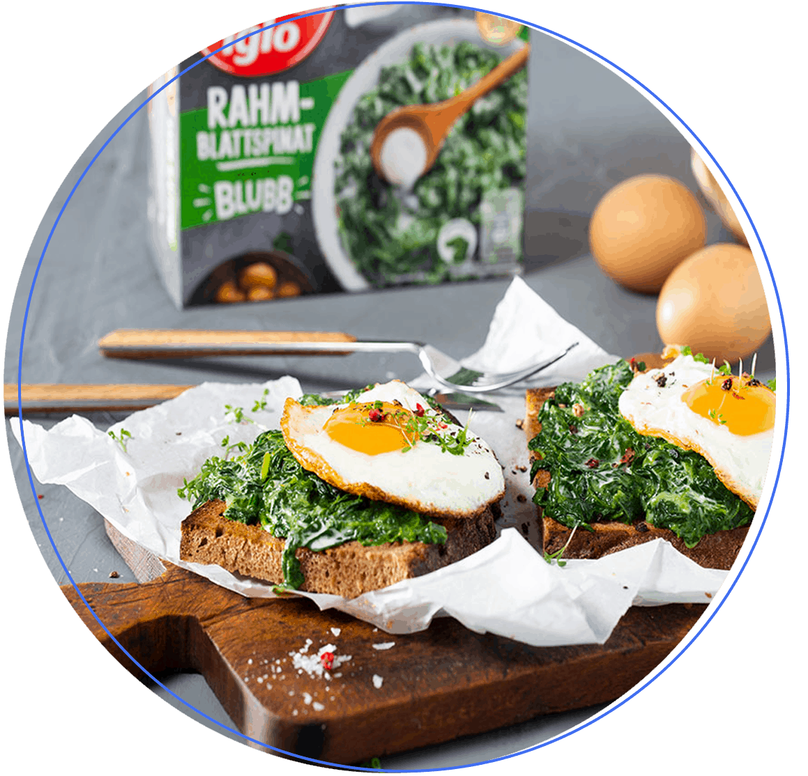 Zwei Scheiben Toast mit cremigem Spinat und Spiegeleiern liegen auf Backpapier auf einem Holzbrett; im Hintergrund stehen frische Eier und eine Schachtel Spinat von Nomad Foods.