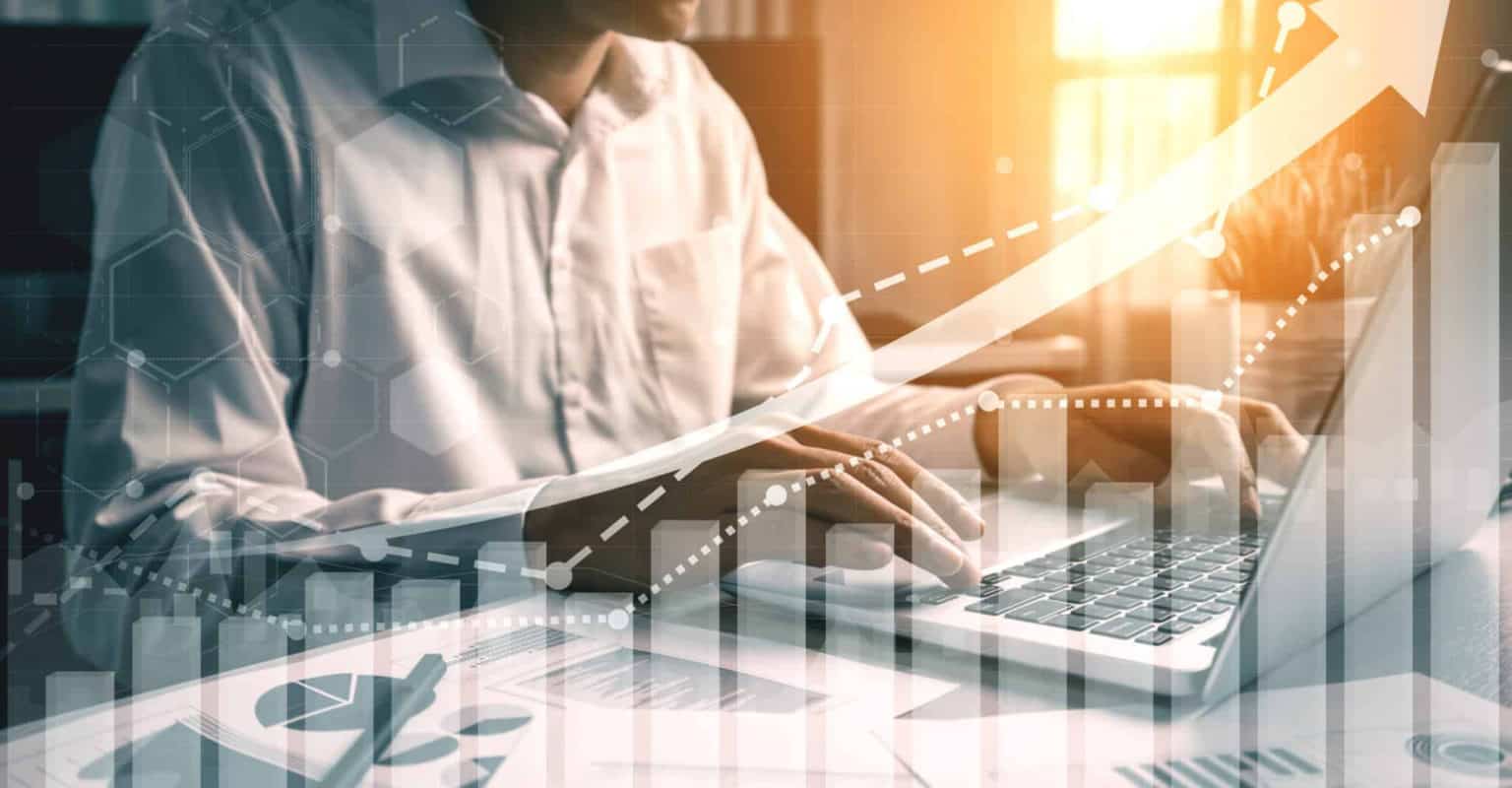 A person in a white shirt types on a laptop with financial charts, graphs, and an upward arrow overlayed, symbolizing business growth and data analysis with Instacart Advertising. Sunlight streams in from the background.