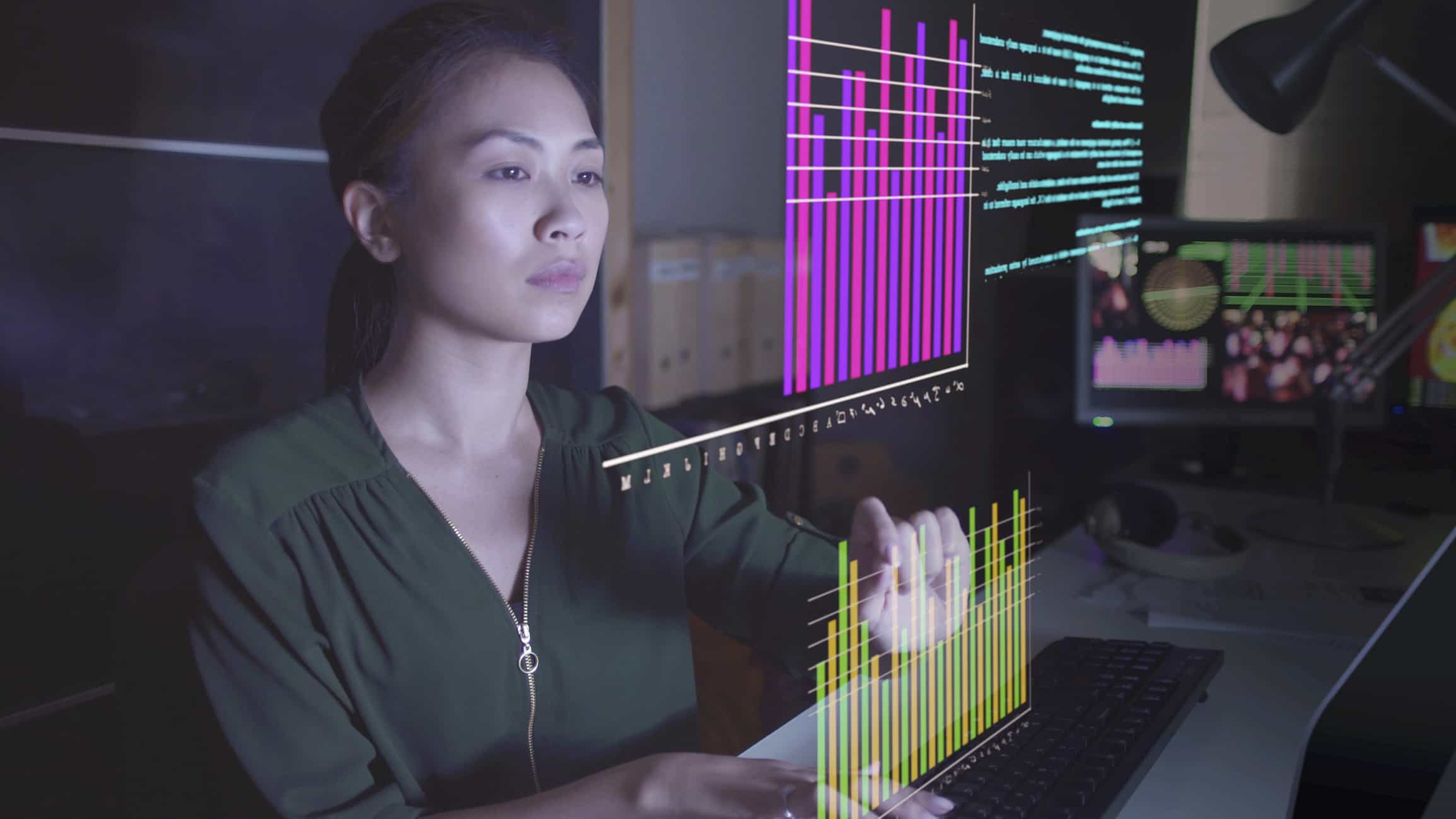 Dans un bureau moderne, une femme est assise à un bureau et tape sur un clavier, tout en interagissant avec des graphiques virtuels colorés et des visualisations de données issues de la modélisation du mix média projetées devant elle.
