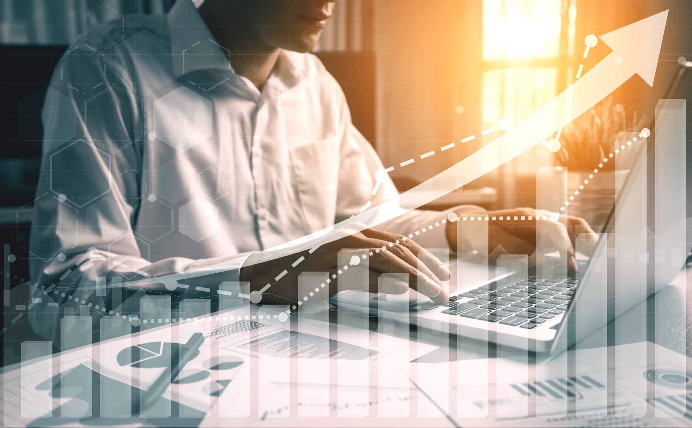 Une personne vêtue d'une chemise blanche travaille sur un ordinateur portable à un bureau où sont disposés des documents, sur lesquels se superposent des graphiques et une flèche pointant vers le haut, symbolisant la croissance de l'entreprise, la réussite financière et l'efficacité de la modélisation du mix média. La lumière du soleil pénètre à flots par une fenêtre.