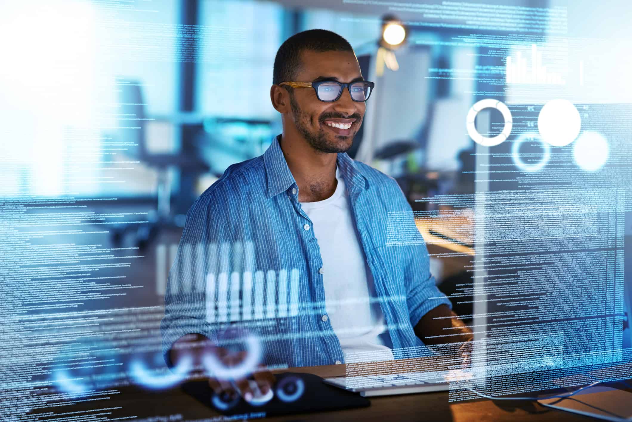 A smiling man wearing glasses and a blue shirt sits at a desk using a computer, surrounded by transparent digital data and graphs, suggesting he is working with technology or media mix modeling.