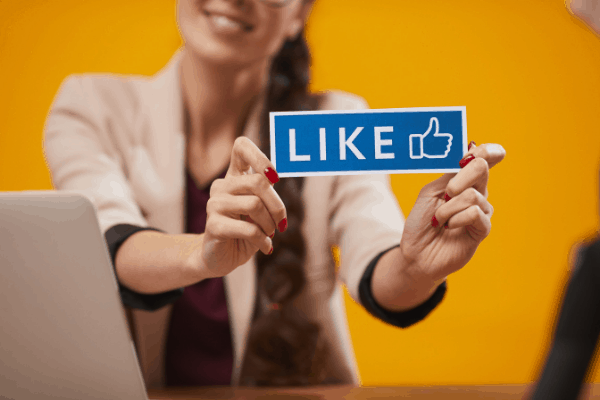 Woman sitting at a desk holding a sign with like written out and a thumbs-up icon.