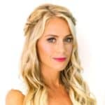 Une femme aux longs cheveux blonds ondulés, coiffés en boucles souples et agrémentés de tresses, portant du rouge à lèvres rose et un haut blanc, pose devant un fond blanc.