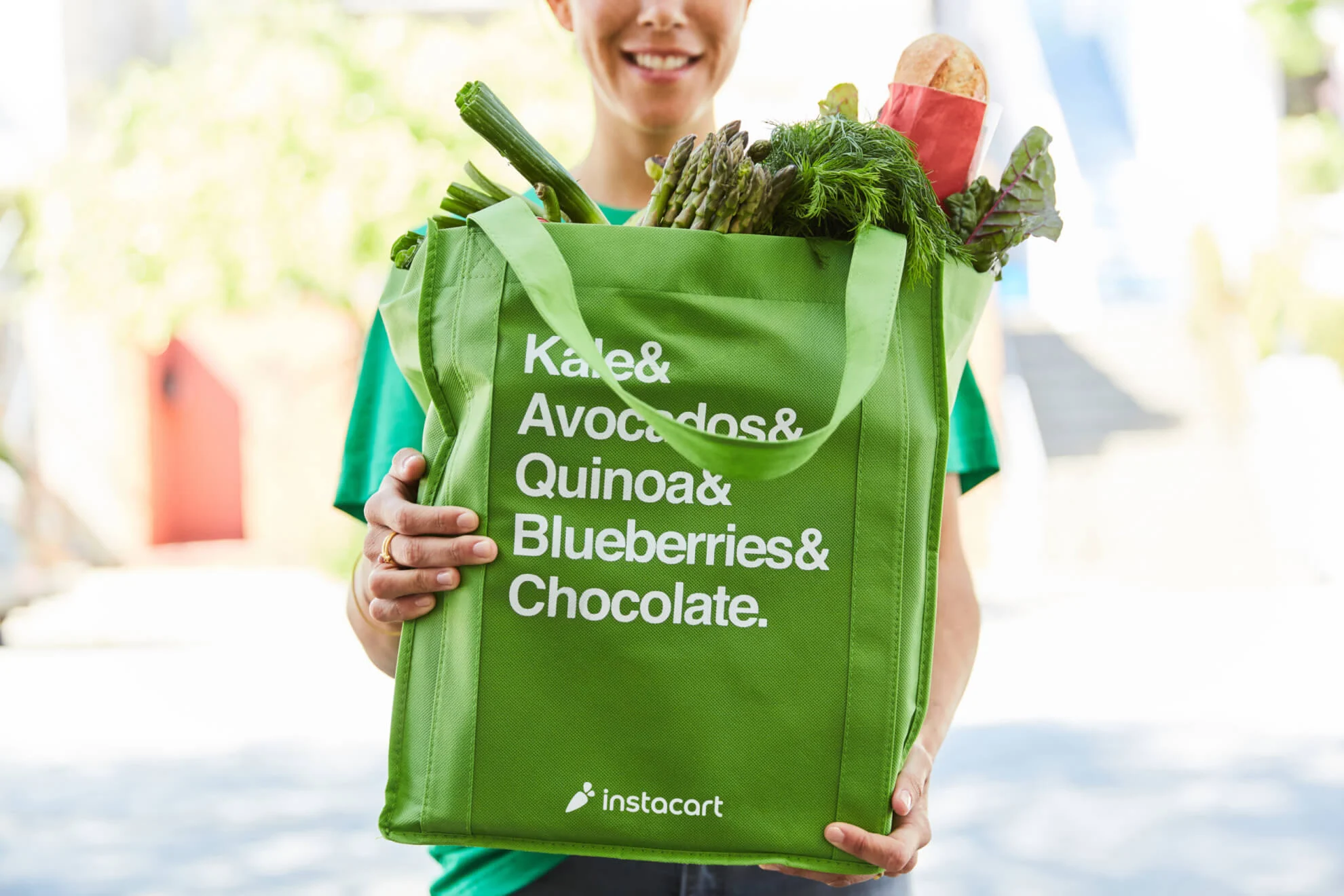 A person holding a green Instacart tote bag filled with groceries like asparagus, bread, and greens promotes Instacart Ads. The bag reads, Kale & Avocados & Quinoa & Blueberries & Chocolate.