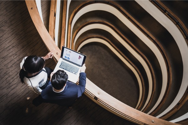 Two business people standing over a tall stairwell while looking at a laptop showing charts