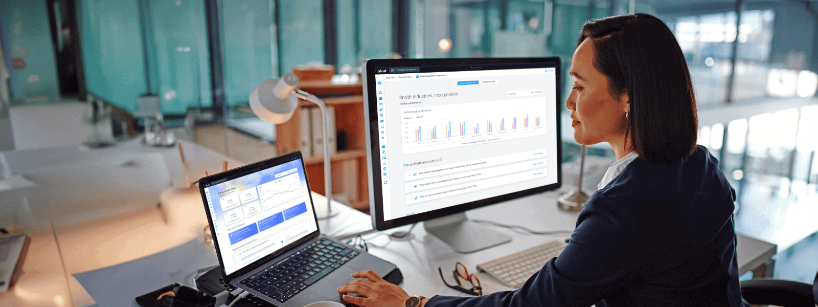 Eine Person nutzt in einem Kreativzentrum einen Laptop, auf dem die Benutzeroberfläche einer Asset-Management-Software zu sehen ist, mit verschiedenen Abbildungen von Geräten, Beschreibungen und Daten in einem Rasterlayout. Im Hintergrund sind Bücher und eine Pflanze zu sehen.