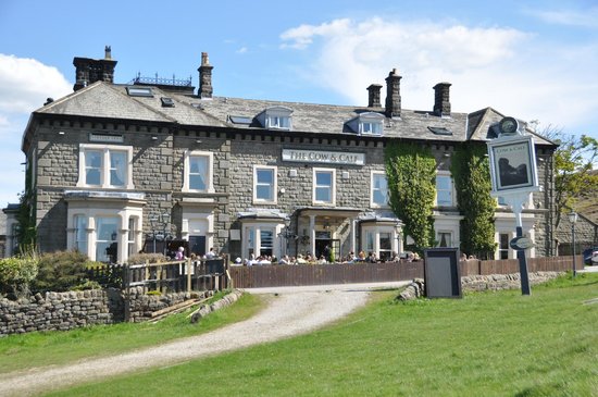 Une grande bâtisse en pierre aux finitions blanches, avec une enseigne indiquant « The Cow & Calf » ; une terrasse extérieure où sont attablés des clients — dont Ben O'Mahoney, d'Ocado Retail —, entourée de pelouse, d'un mur de pierre et d'arbres, par une journée ensoleillée.