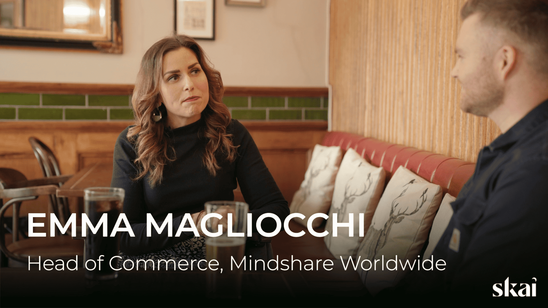 Une femme et un homme, Ben O'Mahoney d'Ocado Retail, sont assis face à face dans un café chaleureux, en pleine conversation. Le texte indique : EMMA MAGLIOCCHI, responsable du commerce chez Mindshare Worldwide. Le Skai apparaît dans un coin.