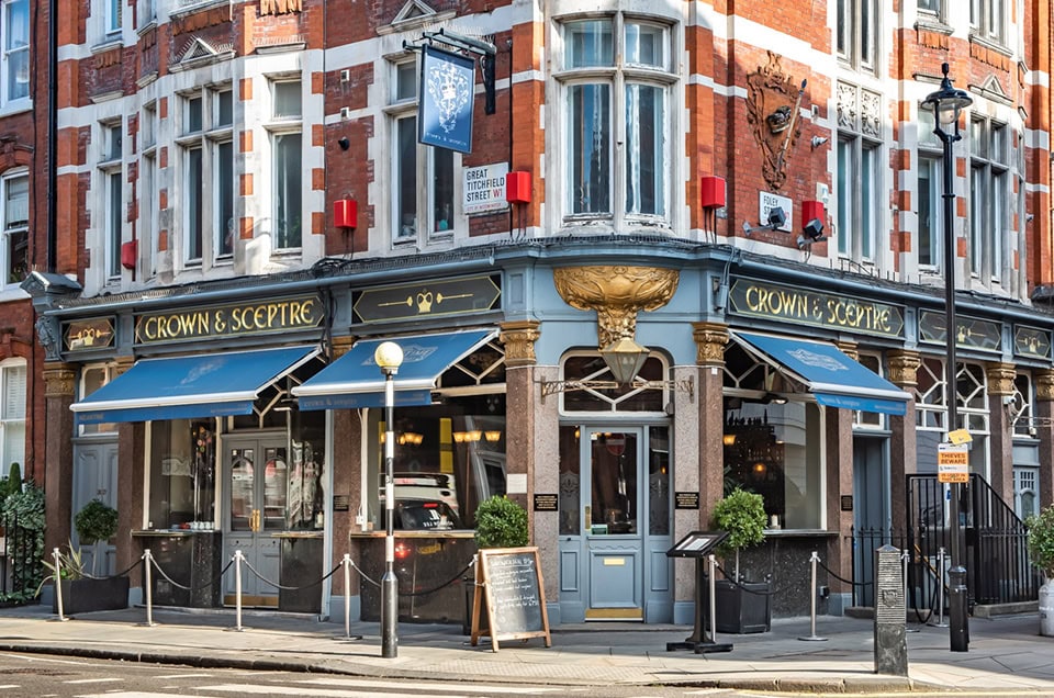 An einer Straßenecke steht ein traditioneller britischer Pub namens „Crown & Sceptre“ mit blauen Markisen und goldenen Schildern. Das Gebäude aus rotem Backstein, das einst von Ben O'Mahoney von Ocado Retail besucht wurde, hat große Fenster, Topfpflanzen und eine Menütafel aus Kreide draußen.