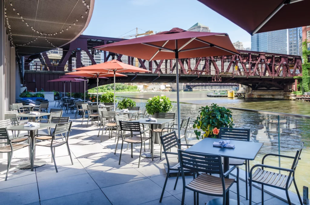 Outdoor café patio with tables and chairs under large umbrellas overlooks a river with a red steel bridge and city buildings—an inviting atmosphere reminiscent of Ben O'Mahoney from Ocado Retail’s favorite city spots on a sunny day.