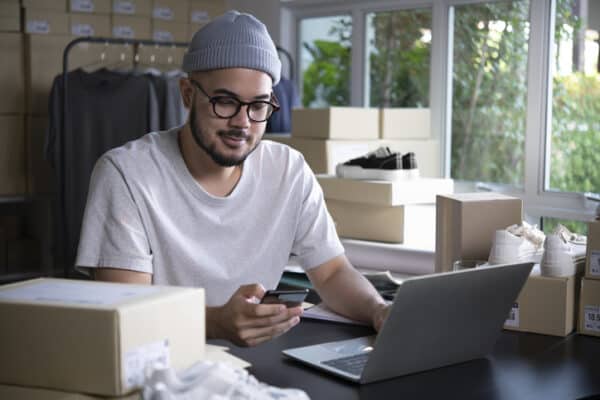 Man using his laptop and smartphone surrounded by boxes and apparel