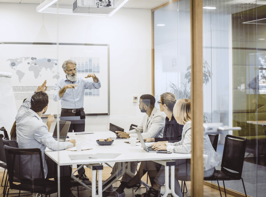 Dans une salle de réunion moderne, un groupe de six professionnels est assis autour d'une table de conférence, ordinateurs portables à la main, et écoute un homme d'un certain âge qui, debout, fait une présentation sur la sécurité des données.