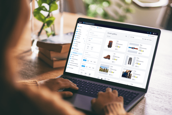 Eine Person sitzt an einem Holztisch und nutzt ihren Laptop, um eine Bilddatenbank mit Produktbildern, Beschreibungen und Bewertungen auf dem Bildschirm zu durchstöbern. Bücher und eine Pflanze im Hintergrund sorgen für eine gemütliche, kreative Atmosphäre.
