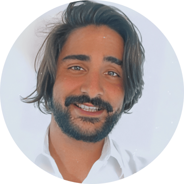 Un homme aux cheveux bruns mi-longs, portant une barbe fournie et une moustache, qui sourit. Il porte une chemise blanche et se détache sur un fond uni de couleur claire, comme on peut le voir dans notre étude de cas sur l'optimisation des destinations.