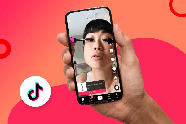 Eine Hand hält ein Smartphone, auf dem ein TikTok-Video-Shopping-Anzeigen-Clip zu sehen ist, in dem sich jemand Mascara aufträgt. Das TikTok-Logo befindet sich in der unteren linken Ecke vor einem bunten Hintergrund mit einem Farbverlauf aus Orange und Rosa sowie kreisförmigen Motiven.