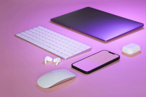 A laptop, wireless keyboard, mouse, smartphone, earbuds, and charging case are arranged neatly on a pink surface under purple lighting—an ideal setup for a consumer electronics advertising case study.