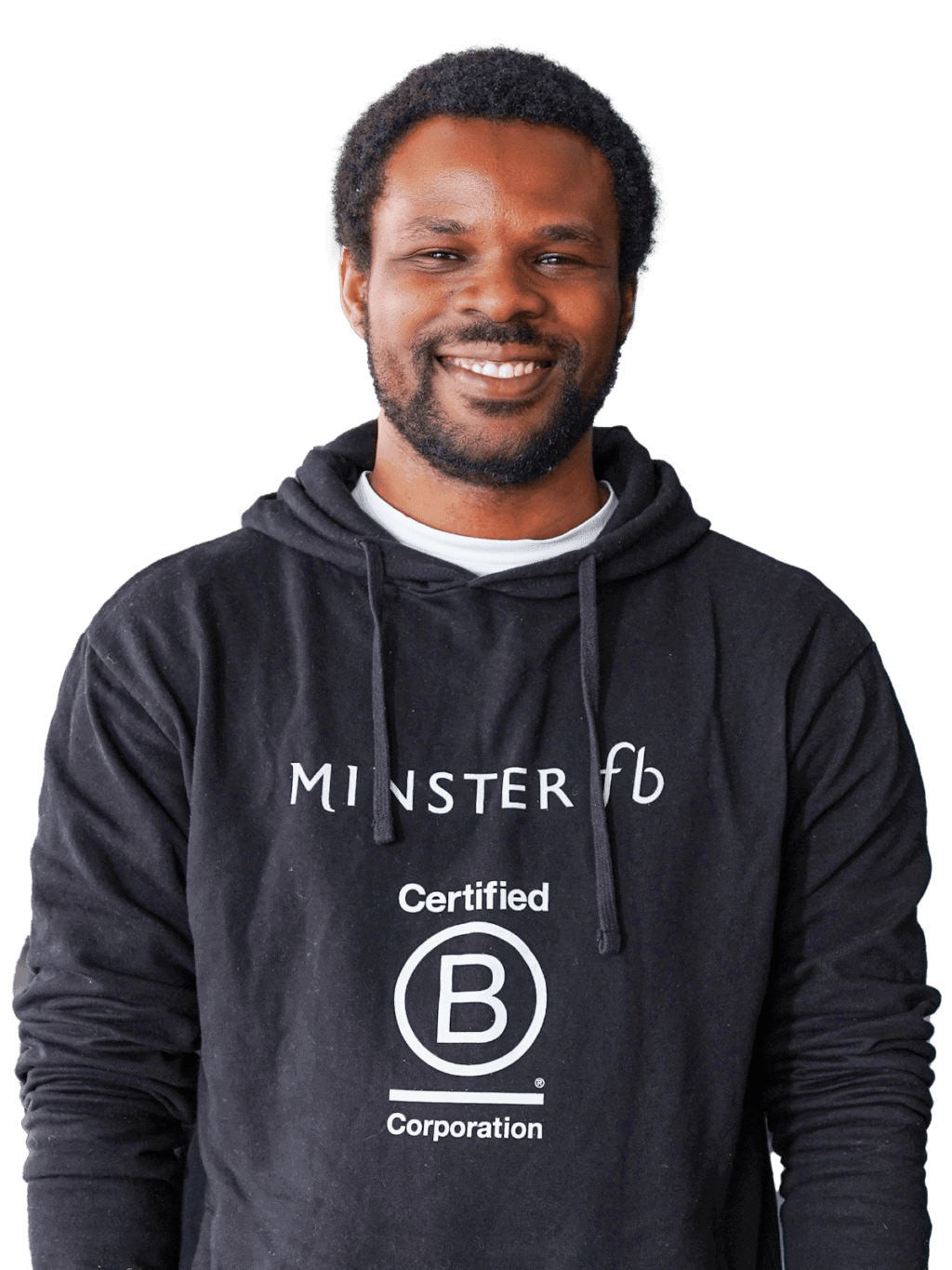 Un homme aux cheveux courts et bouclés, portant une barbe, sourit à la caméra. Il porte un sweat à capuche noir sur lequel sont imprimés les mots « MINSTER fb » et « Certified B Corporation ». L'arrière-plan est d'un blanc uni.