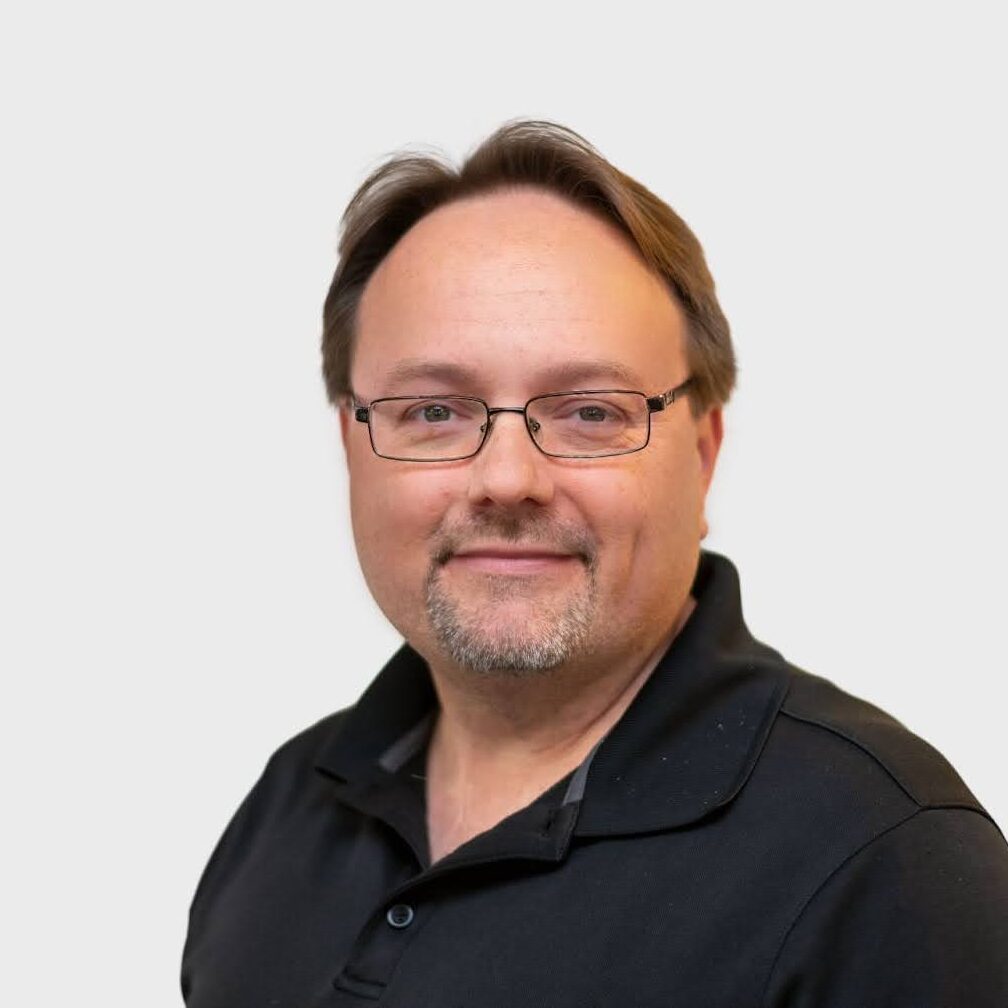 A middle-aged man with short brown hair, glasses, and a trimmed goatee is smiling slightly. He is wearing a black collared shirt and is posed against a plain light gray background.