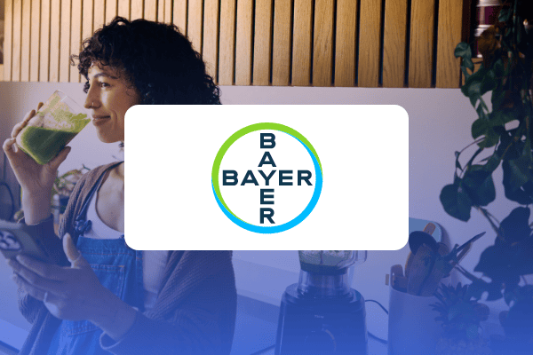Eine Frau in einer Küche trinkt einen grünen Smoothie, während sie ein Handy in der Hand hält. In der Bildmitte ist das Bayer-Logo über dem Bild zu sehen, was eine Fallstudie zu Retail-Media-Werbung in der Praxis verdeutlicht.
