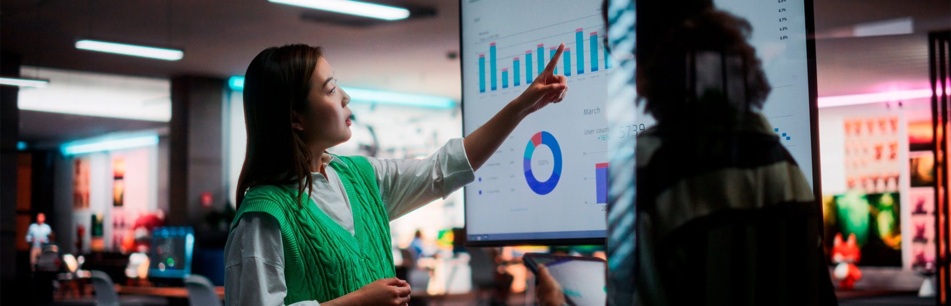 Eine Frau in einer grünen Weste zeigt in einem modernen Büro auf Grafiken und Diagramme auf einem großen Bildschirm und präsentiert einem Kollegen Daten zu den Trends im Bereich Retail Media für das Jahr 2025.