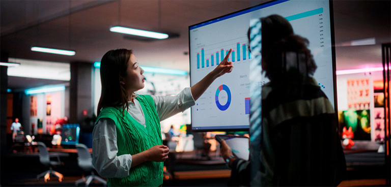 Eine Frau zeigt auf einen digitalen Bildschirm, auf dem Balken- und Kreisdiagramme zu sehen sind, und bespricht mit einem Kollegen in einem modernen, farbenfrohen Büro die Trends im Bereich Retail Media für 2025.