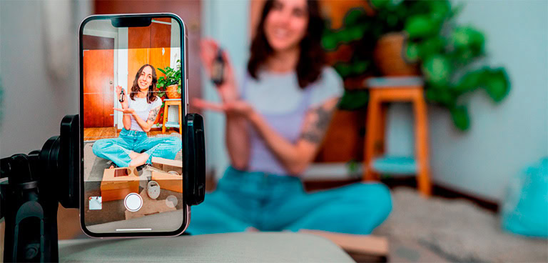 Eine Frau sitzt im Schneidersitz und filmt sich selbst mit einem Smartphone auf einem Stativ, während sie ein Produkt in einem hellen, gemütlichen Raum mit Pflanzen und Holzdekor in die Kamera hält – ein Einblick in die Welt der Retail Media im Jahr 2025, während der Bildschirm ihres Handys ihre Pose scharf hält.