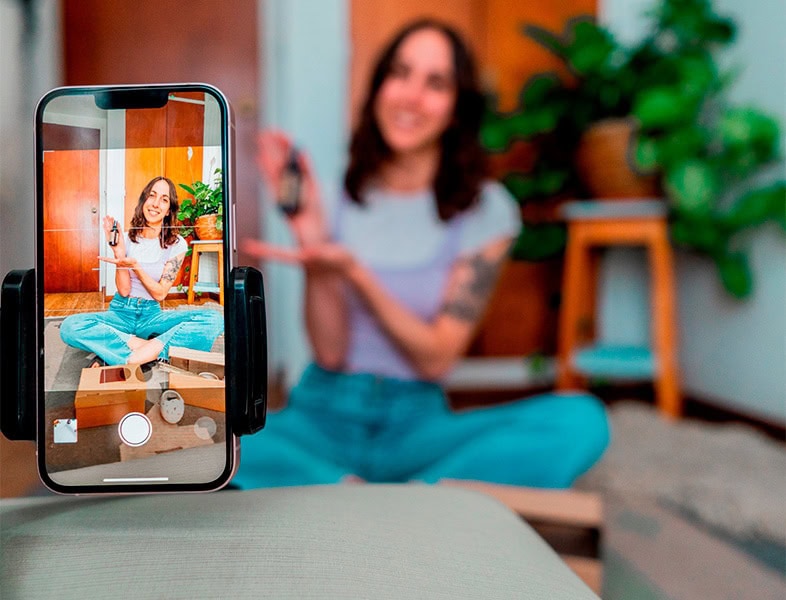 Eine Frau sitzt zu Hause mit gekreuzten Beinen auf dem Boden, lächelt und gestikuliert, während sie über Trends im Bereich Retail Media für 2025 spricht. Das Ganze wird mit einem auf einem Stativ befestigten Smartphone aufgezeichnet. Im Hintergrund sind eine Pflanze und Holzmöbel zu sehen.