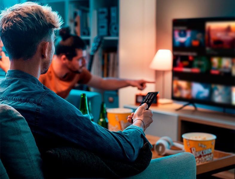 Zwei Leute sitzen auf einer Couch in einem gemütlichen Wohnzimmer und schauen mit der Fernbedienung fern. Der eine zeigt auf den Bildschirm, während der andere die Fernbedienung in der Hand hält und über Retail Media 2025 spricht. Auf dem Tisch stehen Popcorn und Getränke, und im Hintergrund leuchtet eine Lampe.