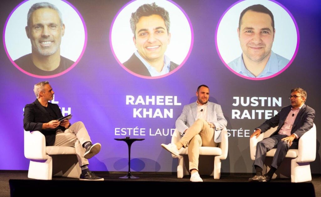 Three men sit on stage in white chairs, engaging in a panel discussion. Behind them is a large screen displaying headshots and names: Joel Heath, Raheel Khan, and Justin Raiten, along with the Estée Lauder logo.