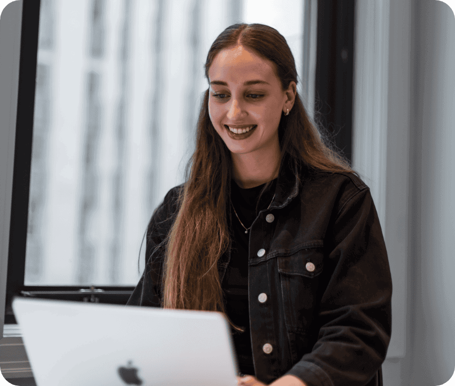 Une jeune femme aux longs cheveux bruns, vêtue d'une veste en jean noire, sourit tout en travaillant sur un ordinateur portable dans un bureau moderne doté de grandes baies vitrées.
