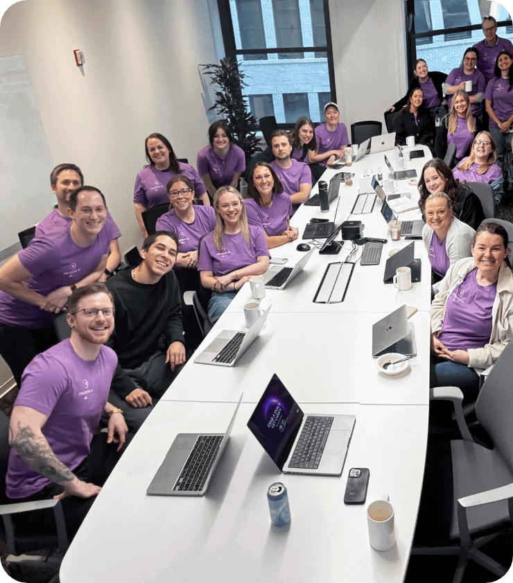 Un groupe d'une vingtaine de personnes, dont beaucoup portent des chemises violettes assorties, sourient et prennent la pose autour d'une longue table de réunion blanche, sur laquelle sont posés des ordinateurs portables, des tasses et des boissons, dans une salle de réunion moderne.