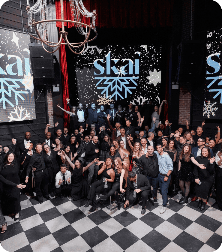 Un grand groupe de personnes pose et sourit ensemble sur un sol à carreaux noirs et blancs, devant une scène où skai sur des écrans, sous des décorations festives en forme de flocons de neige et un lustre.
