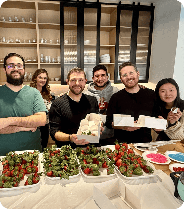 Un groupe de cinq personnes souriantes se tient derrière une table recouverte de plateaux remplis de fraises enrobées de chocolat et de vermicelles. Certaines tiennent des boîtes ouvertes contenant des fraises, et on aperçoit à l'arrière-plan des étagères sur lesquelles sont disposés des verres.