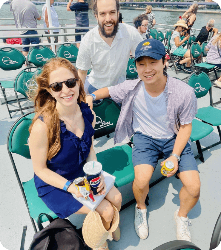 Trois amis sourient, assis sur des sièges verts sur le pont d'un bateau. Une femme portant des lunettes de soleil est assise, un verre et un chapeau à la main, tandis que deux hommes posent à ses côtés. On aperçoit d'autres passagers et l'eau en arrière-plan.