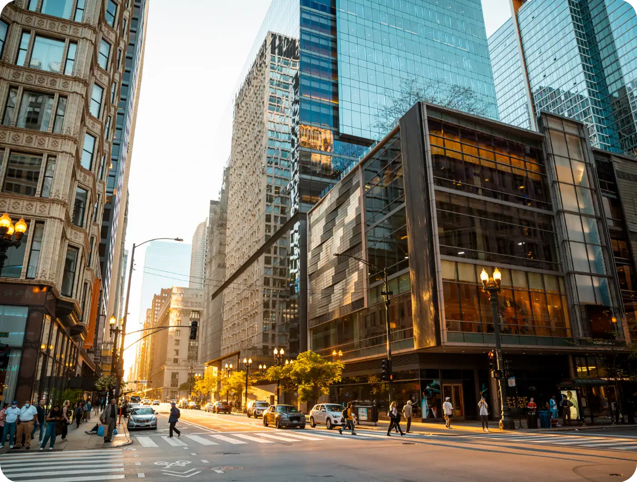 Busy city street intersection at sunset, with people crossing and cars waiting; tall glass and brick buildings reflect the warm sunlight, creating a vibrant urban atmosphere.