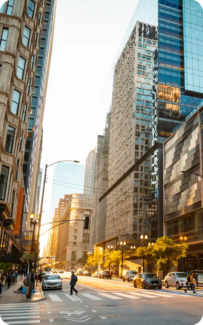 A city street scene at sunset with tall modern glass buildings and older architecture, cars and taxis on the road, and people walking along the sidewalks. Warm sunlight reflects off the buildings.