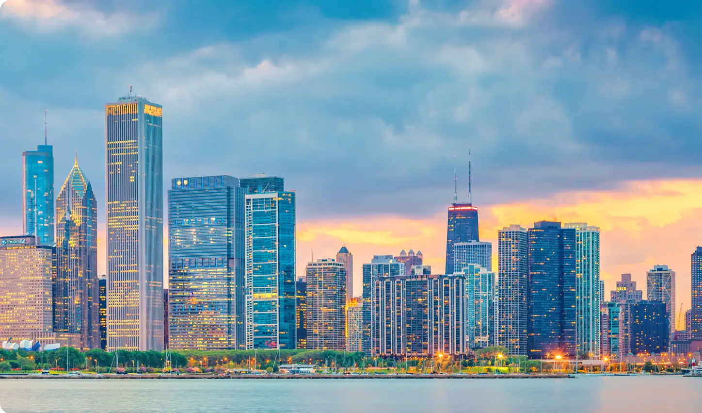 A city skyline with tall buildings and skyscrapers along a waterfront at sunset, with a cloudy sky and reflections on the water.