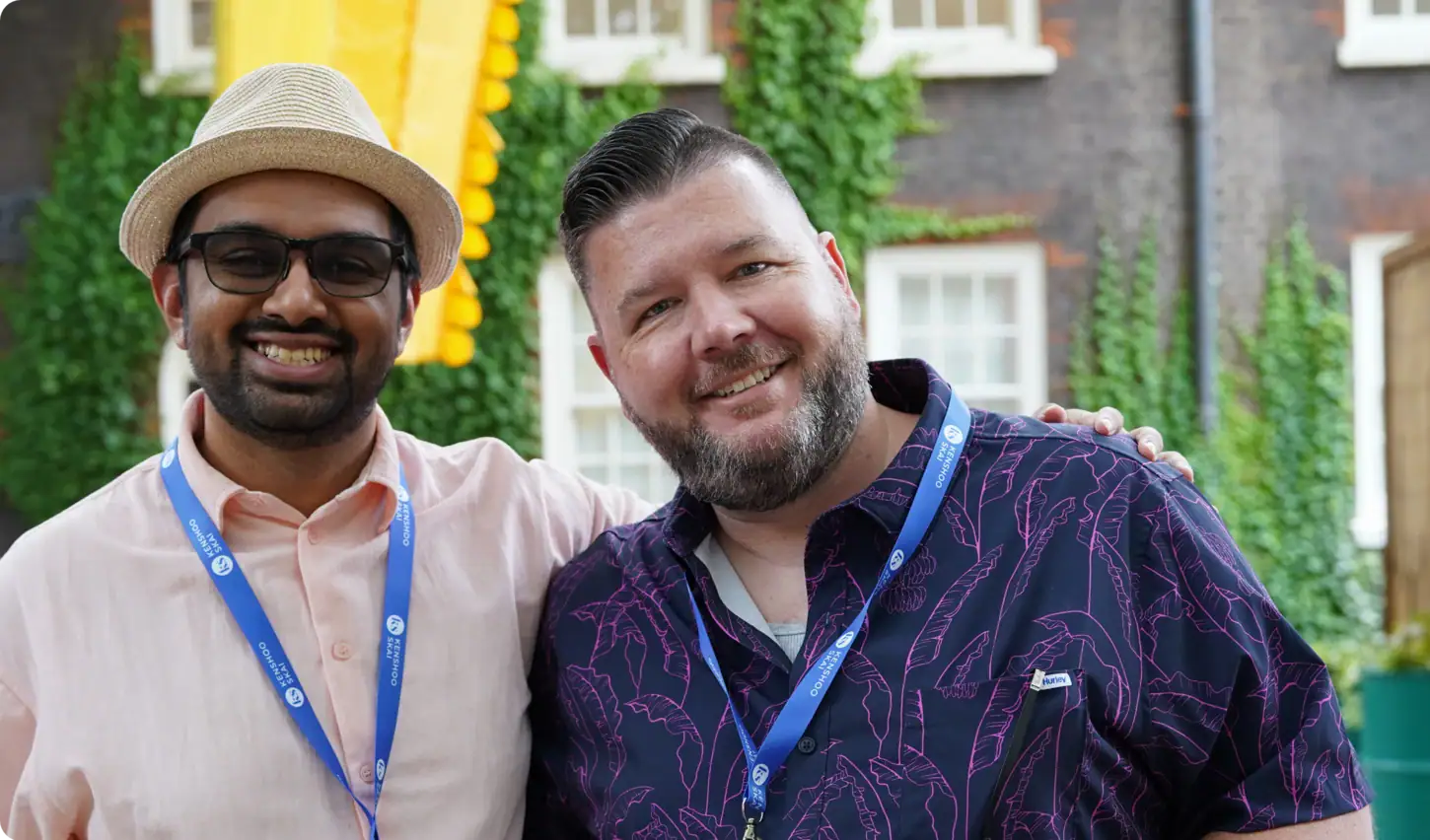 Two men smiling and posing together outdoors, both wearing blue lanyards. One wears a straw hat and glasses, the other has slicked-back hair and a patterned shirt. Greenery and a building with white windows are in the background.
