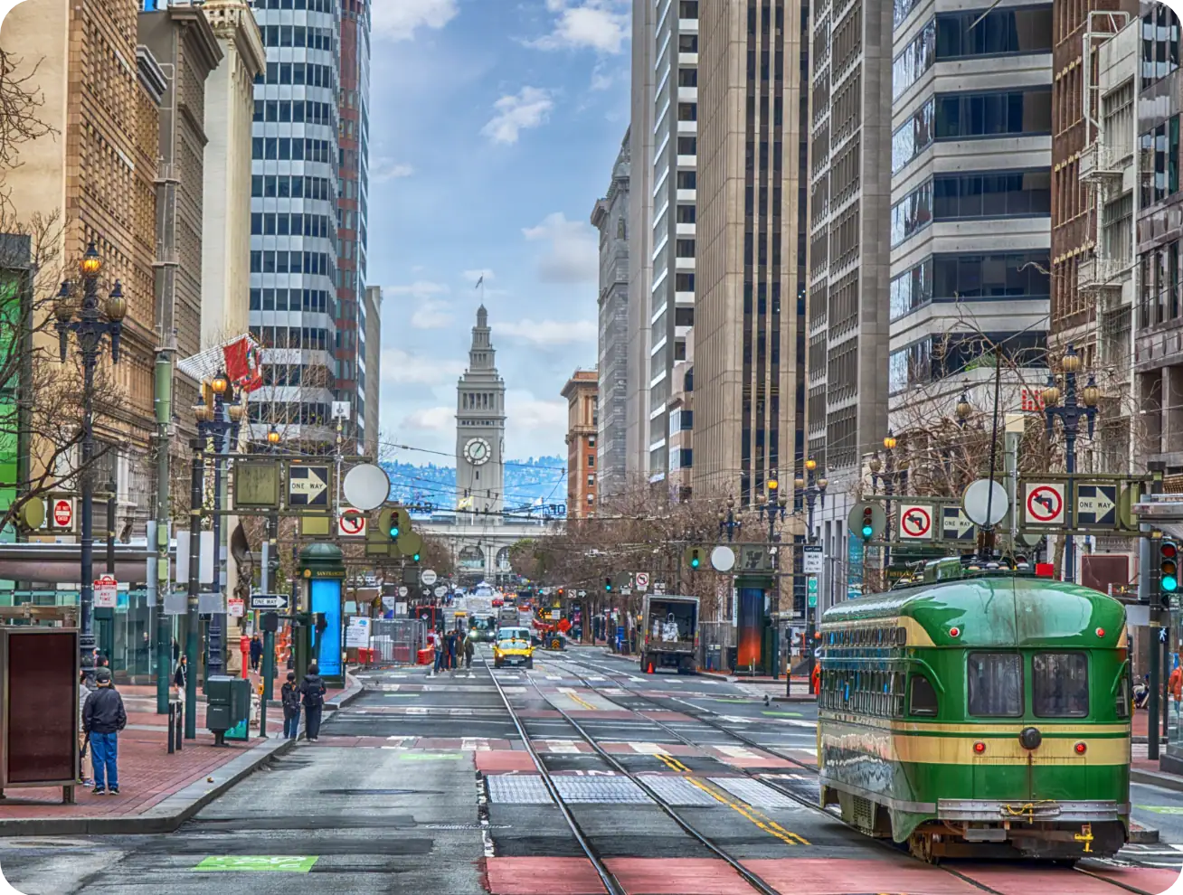 Un tramway vert circule dans une rue de la ville bordée de grands immeubles, avec la tour de l'horloge du Ferry Building visible en arrière-plan sous un ciel partiellement nuageux.