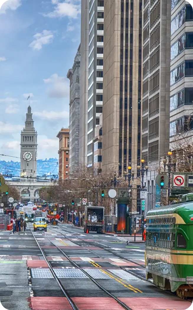 Un tramway circule dans une rue animée de la ville, bordée de grands immeubles, avec, en arrière-plan, le Ferry Building de San Francisco et sa tour horloge, sous un ciel partiellement nuageux.