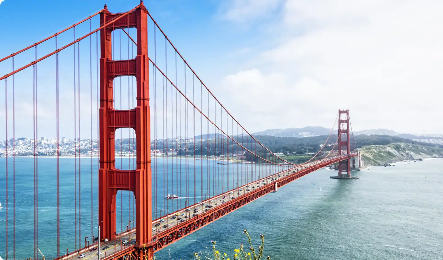 Le Golden Gate Bridge, à San Francisco, enjambe les eaux bleues ; on y voit des voitures qui le traversent, tandis qu'en arrière-plan, sous un ciel partiellement nuageux, se dessinent les immeubles de la ville et les collines.