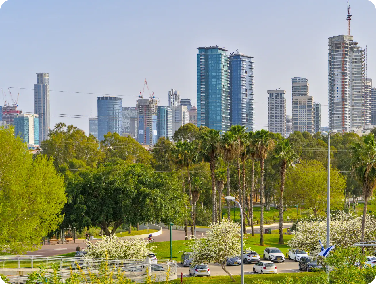 Ein Stadtbild mit modernen Hochhäusern im Hintergrund, üppig grünen Bäumen und Palmen im Vordergrund, einem Park mit Wegen und mehreren geparkten Autos entlang der Straße an einem sonnigen Tag.