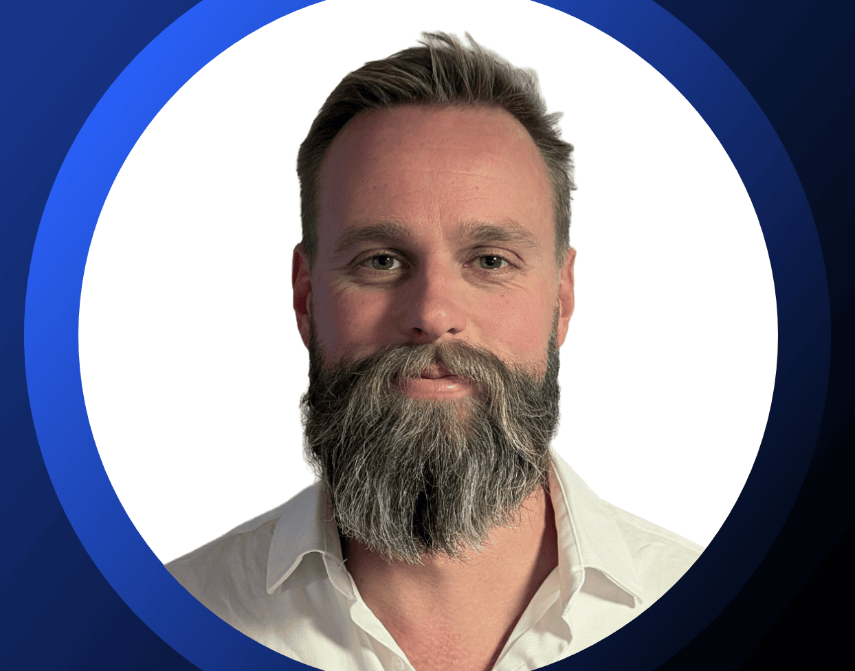 Un homme à la barbe grise fournie et aux cheveux bruns soigneusement coiffés, vêtu d'une chemise blanche à col, pose devant un fond blanc, l'image étant encadrée d'un liseré circulaire bleu.