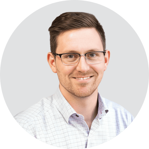 Un homme souriant, aux cheveux bruns courts, portant des lunettes et une chemise à carreaux de couleur claire, pose devant un fond uni gris clair, prêt à partager ses réflexions sur les prévisions concernant le retail media en 2026.