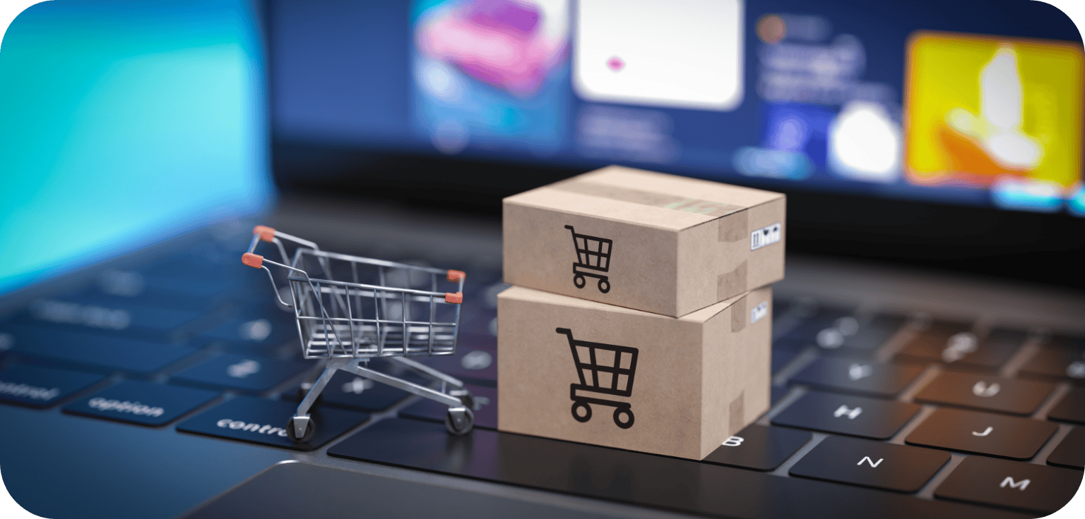 Zwei kleine Pappkartons mit Einkaufswagen-Symbolen und ein Miniatur-Einkaufswagen stehen auf einer Laptop-Tastatur und stehen für Online-Shopping, E-Commerce und die Zukunft der Retail-Medien im Jahr 2026. Im Hintergrund ist ein verschwommener Bildschirm zu sehen.