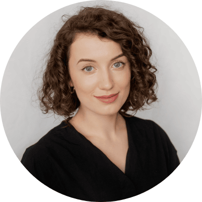 Portrait d'une jeune femme aux cheveux bruns courts et bouclés, au teint clair et aux yeux bleus, vêtue d'un haut noir, souriant doucement à l'appareil photo — idéal pour une étude de cas sur le retail media, sur un fond clair et neutre.