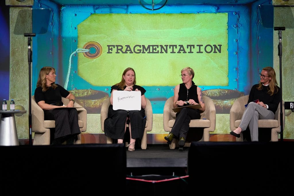 Four women sit on a stage in armchairs, engaged in a panel discussion. One holds up a sign that says Fragmentation. The stage backdrop displays the word FRAGMENTATION in large letters.